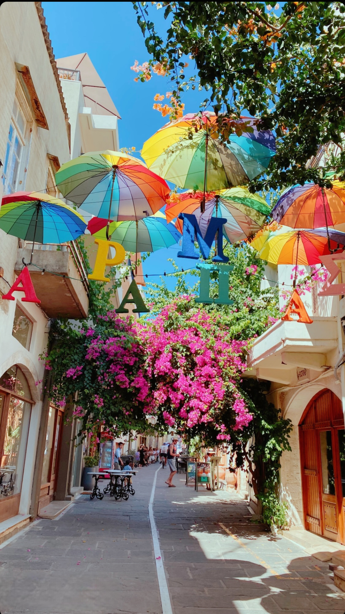 Decorations of multicolor umbrellas and greek writing by pink flowers on a street in Old Town Rethymno